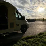 Witte camper met deur open met op de achtergrond een wolkenlucht en een ondergaande zon